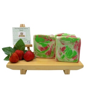 May include: Three bars of handmade soap on a wooden display stand. The soap is green and red with a marbled swirl pattern. The soap is labeled "Strawberry Patch Soap". There are three strawberries and green leaves on the stand.