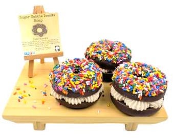 May include: Three donut-shaped soaps with chocolate frosting and colorful sprinkles sit on a wooden tray. A small easel displays a sign that reads "Sugar Cookie Donuts Soap". The soaps have a cream-colored filling.