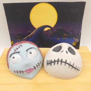 May include: Two bath bombs shaped like characters from The Nightmare Before Christmas. One is light blue with stitched details and pink lips, the other is white with a stitched mouth and eye sockets. A backdrop features a full moon and a spooky landscape.