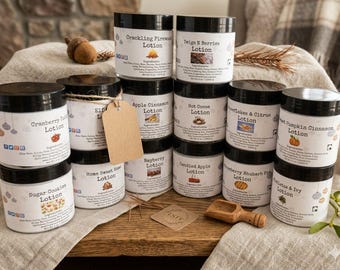 May include: A collection of twelve cylindrical lotion jars with black lids, each featuring a white label with a unique scent name, ingredient list, and small graphic. A blank brown paper tag hangs from one jar, and a small wooden scoop sits nearby.