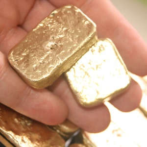May include: A hand holding a stack of gold bars. The bars are rectangular and have a rough, textured surface. The bars are stacked on top of each other, with the top bar being the largest.