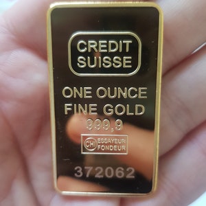 May include: A gold bar inscribed with "CREDIT SUISSE" and "ONE OUNCE FINE GOLD 999.9". The bar also features a stamp reading "ESSAYEUR FONDEUR" and the number "372062".