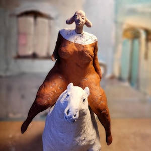 May include: A ceramic sculpture of a woman riding a white horse. The woman is wearing a brown dress and has a white collar. The sculpture is painted in shades of brown, white, and grey.