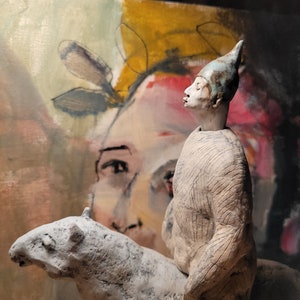 May include: A ceramic sculpture of a person with a pointed hat riding a stylised horse. The sculpture is painted in shades of grey and white, and the background is a colourful abstract painting.