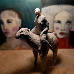 May include: A white ceramic sculpture of a person riding a horse. The sculpture is in a minimalist style with a simple, abstract design. The person is depicted with a white face and a long, flowing robe. The horse is depicted with a long, flowing mane and tail.