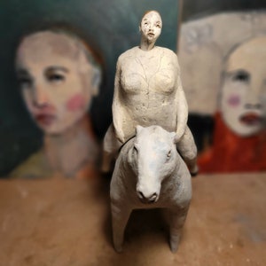 May include: A white ceramic sculpture of a person riding a white horse. The sculpture is in a minimalist style with simple lines and forms.