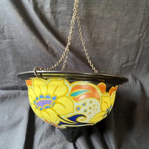 May include: A hanging bowl with a black exterior and interior rim, featuring a vibrant hand-painted floral pattern in yellow, orange, blue, and purple. The bowl is suspended by a thin metal chain attached to the rim, creating a rounded shape.