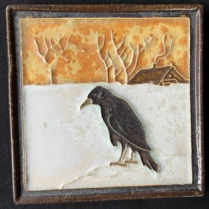 May include: A square ceramic tile featuring a relief design of a black crow standing on a white snowy surface. The background depicts a golden-brown landscape with bare trees and a small house silhouette under a textured, warm-toned sky.