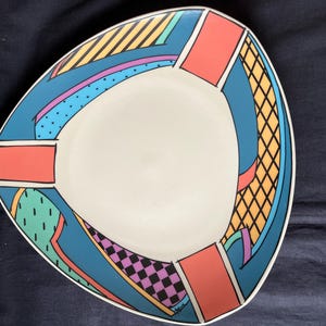 Op de afbeelding: Een driehoekig keramieken bord met een crèmekleurig midden en een brede rand versierd met geometrische patronen in blauw, koraal, geel, groen en paars. Het ontwerp bevat strepen, rasters, stippen en dambordpatronen.