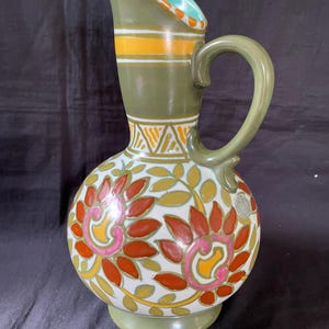 Op de afbeelding: Decoratieve keramische kan met een bolle basis en een slanke hals. De kan heeft een groene handgreep en tuit, met oranje en gele strepen en bloemmotieven in rood, roze en geel. De kan is ongeveer 25 cm hoog.