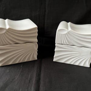 Puede incluir: Dos cajas decorativas de cerámica blanca con un diseño en forma de ola. Cada caja es rectangular con una parte superior curvada y una sección inferior con un patrón radiante. Las cajas están apiladas, creando una apariencia texturizada única.
