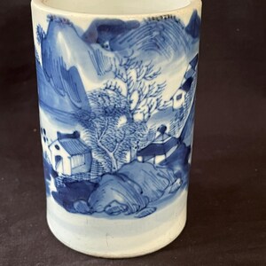 Può includere: Un vaso cilindrico in ceramica bianca con un design a paesaggio blu e bianco. Il vaso presenta una scena di montagna con alberi, case e un fiume.