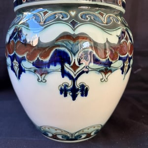 May include: A ceramic vase with a rounded body and a wide rim. The surface features a glossy white base decorated with intricate, symmetrical patterns in shades of deep blue, teal, and reddish-brown, accented by small circular details.