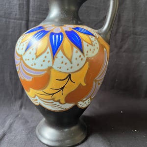 Può includere: Una brocca in ceramica nero opaco con un motivo floreale dipinto a mano in tonalità vivaci di blu, giallo, arancione e viola chiaro. Il recipiente presenta un corpo arrotondato, un collo stretto, un manico curvo e una base a piedistallo.