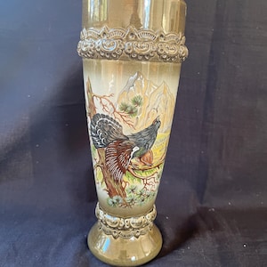 Könnte beinhalten: Ein hoher, brauner Keramik-Bierkrug mit einem detaillierten Design. Der Krug zeigt ein gemaltes Bild eines Auerhuhns, das auf einem Ast in einer Berglandschaft sitzt. Der Krug hat einen dekorativen Rand und Boden.