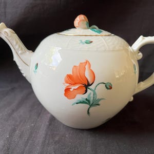 May include: A cream-coloured porcelain teapot featuring a hand-painted orange poppy flower on the side. The lid is topped with a small sculpted flower bud handle. The upper portion of the teapot has a textured basket-weave pattern with gold trim.