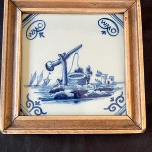 以下が含まれることがあります： 木製の額縁に入った青と白のデルフトタイル。風車と井戸、帆船2隻が描かれている。タイルの右上に「WMO」のイニシャルがある。