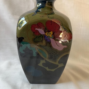 Puede incluir: Un jarrón de cerámica con acabado brillante, que presenta una base oscura que se transforma en una sección superior de color verde oliva. La superficie está decorada con un patrón floral estilizado en tonos rojo, rosa, amarillo y verde azulado, con contornos negros.