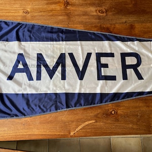 Op de afbeelding: Een marineblauwe en witte wimpel met het woord "AMVER" in marineblauwe hoofdletters. De vlag heeft een witte horizontale streep en is gemaakt van een glanzend materiaal. Hij ligt op een houten oppervlak.
