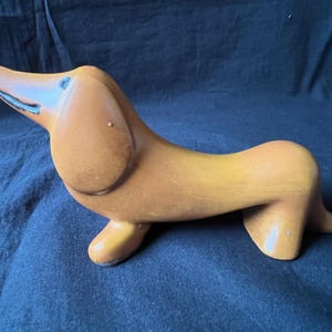 May include: A ceramic dachshund figurine in shades of brown and yellow. The dog is posed with its head up and its long body curved. The figurine has a glossy finish and is set against a dark blue background.