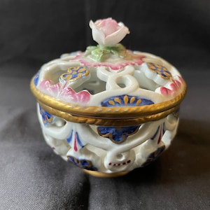 Könnte beinhalten: Eine weiße Porzellan-Schmuckdose mit goldener Verzierung und filigranen Blumendesigns in Blau, Rosa und Gold. Der Deckel ist mit einer zarten rosa Rose verziert.