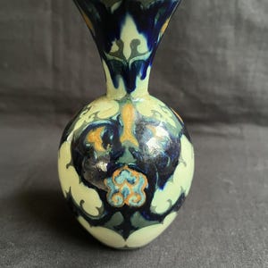 Può includere: Un piccolo vaso in ceramica bianca con corpo rotondo e collo stretto. Il vaso è decorato con intricati motivi floreali blu, verdi e dorati.