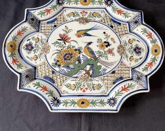 Könnte beinhalten: Ein weißer Keramikteller mit einem blauen, gelben und grünen Blumendesign und zwei Vögeln. Der Teller hat einen gewellten Rand und ein geometrisches Muster in der Mitte.