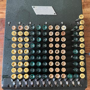 Puede incluir: Una calculadora mecánica Comptometer vintage de color verde oscuro con una cuadrícula de teclas numeradas en amarillo, beige y verde oscuro. Una placa de metal en la parte superior muestra el texto Comptometer. El dispositivo incluye varias columnas.