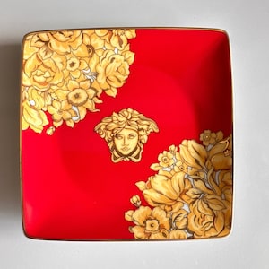 Puede incluir: Bandeja cuadrada de cerámica roja con un diseño floral dorado y una cabeza de Medusa dorada en el centro.