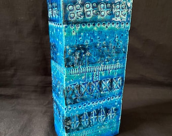 May include: A tall, rectangular ceramic vase with a blue glaze and intricate geometric patterns. The vase has a textured surface and a slightly iridescent finish.