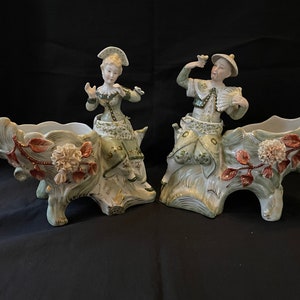 Peut inclure: Une paire de figurines en porcelaine représentant un planteur, chacune avec une personne en costume coloré assise sur une souche d'arbre avec une fleur et des feuilles. Les planteurs sont blancs avec des accents verts et dorés.