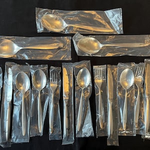 May include: A set of new, silver-coloured cutlery, including spoons, forks, and knives, individually wrapped in clear plastic. The utensils are arranged on a black surface, ready for use.