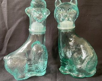 Peut inclure: Deux bouteilles en verre en forme de caniche et de chat. Les bouteilles sont de couleur bleu-vert clair et ont une surface texturée. Les bouteilles ont des bouchons en verre transparent.