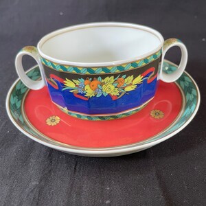 May include: A red and white porcelain soup bowl with a blue and gold floral design. The bowl has two handles and is sitting on a matching saucer.