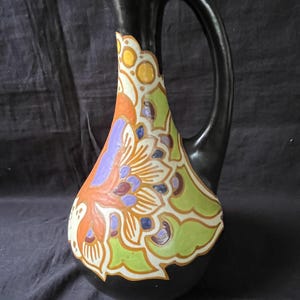 Può includere: Brocca in ceramica nera con manico curvo e beccuccio. La brocca è decorata con un colorato motivo floreale in arancione, verde, viola e giallo su sfondo bianco.