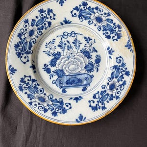 Puede incluir: Un plato de Delftware azul y blanco con un diseño floral. El plato tiene un borde amarillo y un motivo floral central en un jarrón.