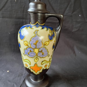 May include: A brown ceramic pitcher with a lid and handle. The pitcher has a cream-colored body with blue, purple, yellow, and orange floral designs. The base and lid are dark brown. The pitcher is likely vintage or antique.