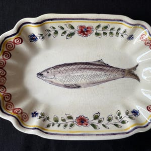 Pode incluir: Uma travessa de servir oval em cerâmica com base cor de creme e acabamento de esmalte craquelado. O centro apresenta uma ilustração detalhada de um peixe. A borda recortada é decorada com um padrão repetitivo de espirais vermelhas e flores.