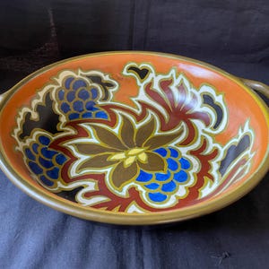 Könnte beinhalten: Ovale Keramik-Servierschüssel mit Griffen. Die Schüssel hat einen leuchtend orangefarbenen Hintergrund mit einem Blumendesign in Braun, Blau, Rot und Gelb. Der Rand und die Griffe sind in gedämpftem Gold gehalten.