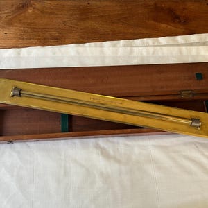 Puede incluir: Un instrumento antiguo de latón y madera, posiblemente una herramienta de medición, descansa en un estuche de madera abierto. El componente de latón es rectangular con una varilla central y accesorios en los extremos. El estuche es marrón oscuro con forro de fieltro verde.