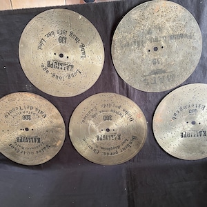 Könnte beinhalten: Fünf antike Musikplatten mit Löchern für eine Spieluhr. Die Platten sind aus Metall und haben einen abgenutzten, alten Look. Die Platten haben Text darauf, darunter "Long Long Ago", "Vals del Teвого", "Dich", "Benheur" und "KALLIGPR".