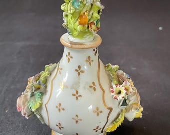 May include: A white porcelain perfume bottle with a floral design. The bottle has a gold trim and a floral stopper. The bottle is decorated with flowers in shades of pink, yellow, green, and blue.