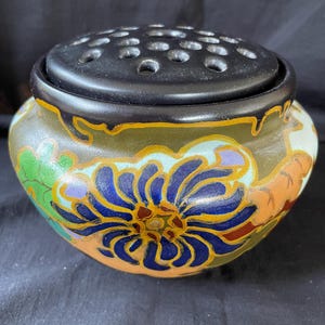 May include: A ceramic flower frog with a black lid featuring multiple holes. The bowl is decorated with a large blue floral design, with accents of green, orange, and yellow. The rim is outlined in black.