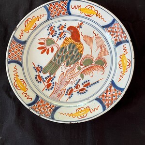 Puede incluir: Un plato de cerámica blanca con un diseño pintado a mano. El plato presenta un pájaro colorido posado en una rama, rodeado de patrones florales y geométricos en rojo, azul y amarillo. El plato tiene un diámetro de aproximadamente 23 cm.