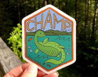 Op de afbeelding: Een zeshoekige sticker met een cartoonillustratie van Champ, het meermonster van Lake Champlain, Vermont. De sticker is blauw, groen en oranje met de tekst "Champ EST. in VT" en "@WANDERONWORDS".