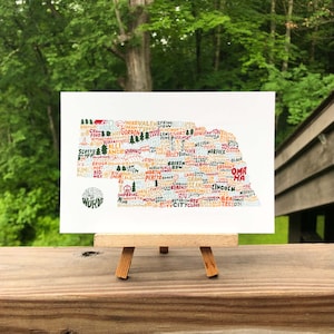 May include: A white rectangular print on a small wooden easel. The print features a colorful map of a state, composed of various city names. The words "Wander and Words" are printed on the lower left.