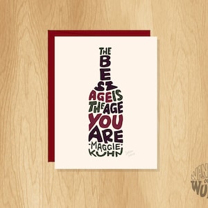 May include: Greeting card with a burgundy border and a cream-colored center. The card features a wine bottle shape filled with the words "THE BEST AGE IS THE AGE YOU ARE - MAGGIE KUHN" in various colors. The words "WANDER ON WORDS" are in a circle in the bottom right.