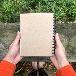 Puede incluir: Un cuaderno con espiral y cubierta de papel kraft se muestra abierto. Las p&aacute;ginas son blancas y la encuadernaci&oacute;n en espiral es negra. El fondo incluye una superficie de madera y vegetaci&oacute;n.