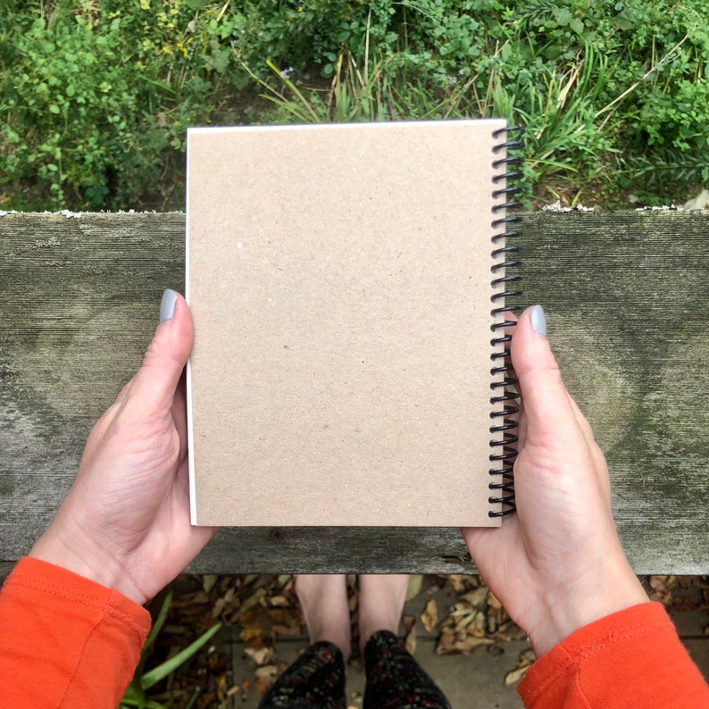 Puede incluir: Un cuaderno con espiral y cubierta de papel kraft se muestra abierto. Las p&aacute;ginas son blancas y la encuadernaci&oacute;n en espiral es negra. El fondo incluye una superficie de madera y vegetaci&oacute;n.