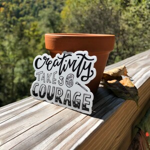 May include: White sticker with black text that reads "Creativity takes courage" with a small circle that says "Henry Matisse" in the middle. The sticker is on a wooden surface next to a terracotta pot.